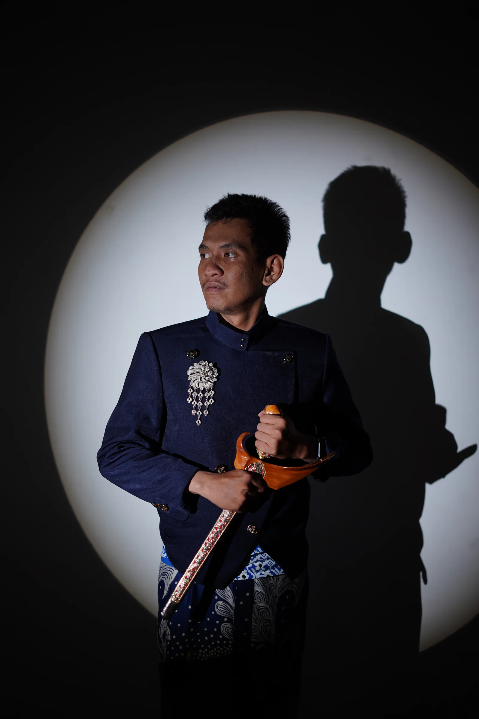 Fadli berdiri dengan keris dalam busana navy dan bayangan yang tegas