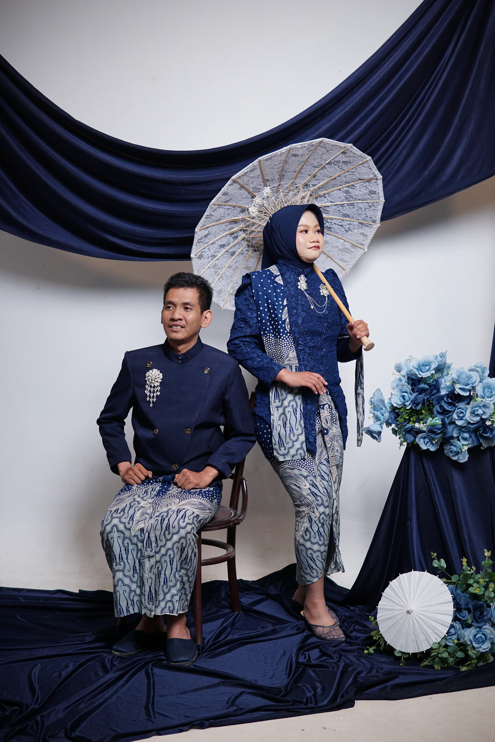 Dewi berdiri dengan payung renda sementara Fadli duduk di sampingnya